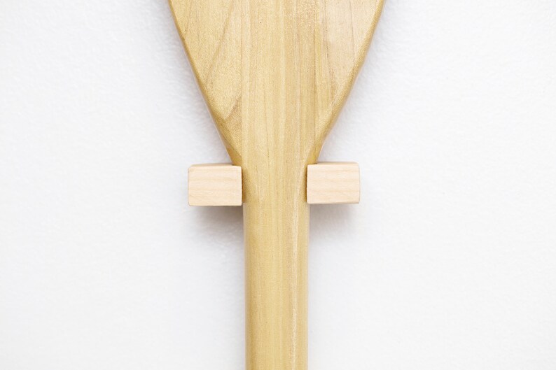 May include: A wooden paddle with a light brown finish is mounted on a white wall using two small wooden blocks.