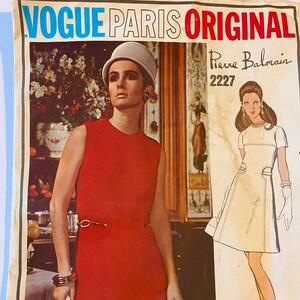 May include: A vintage Vogue Paris Original sewing pattern featuring a red dress design by Pierre Balmain. The pattern number is 2227.