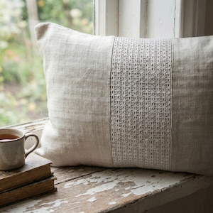 Op de afbeelding: Een rechthoekig beige linnen kussen met een decoratief wit kanten paneel in het midden. Het kussen ligt op een verweerde witte houten vensterbank. Een kopje thee en boeken staan op de voorgrond.