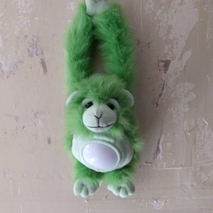 May include: A plush green monkey toy with a white belly, hanging from its arms. The monkey has a furry texture and a smiling face. The toy is hanging against a neutral-colored wall.