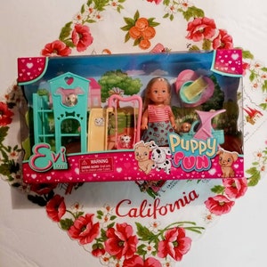 May include: A boxed Evi Love Puppy Fun playset featuring a doll, puppies, and a playground. The packaging is pink and teal, with the words "Puppy Fun" and "Evi Love" displayed. The set includes a swing set and a dog house.