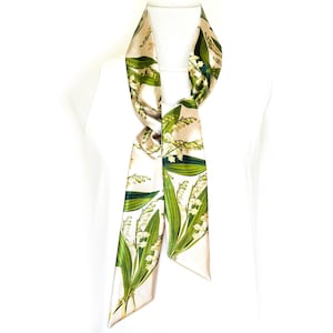 May include: A cream-colored silk scarf with a botanical print of lily of the valley flowers and leaves in green and white. The scarf is long and narrow, designed to be worn around the neck or as an accessory.