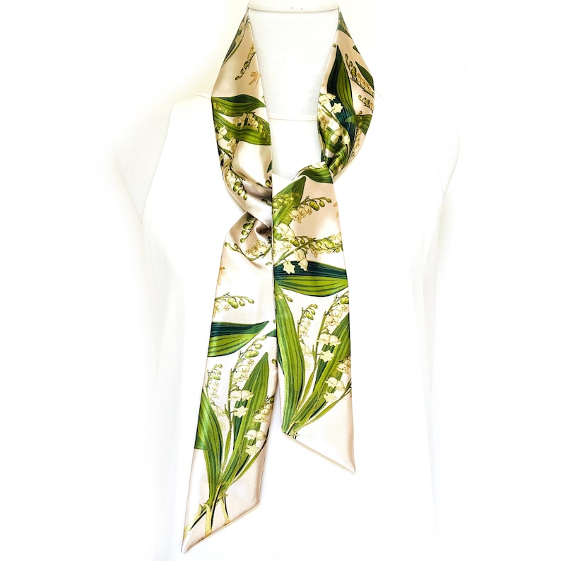 May include: A cream-colored silk scarf with a botanical print of lily of the valley flowers and leaves in green and white. The scarf is long and narrow, designed to be worn around the neck or as an accessory.
