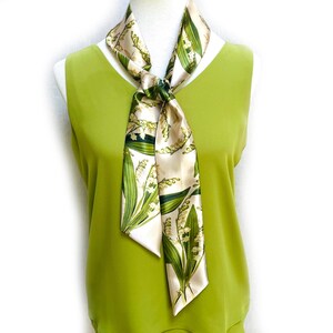 May include: A cream-colored silk scarf with a botanical lily of the valley print. The scarf is tied in a knot, with the ends hanging down. The scarf has green leaves and white flowers. The scarf is draped over a green top.