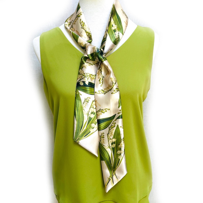 May include: A cream-colored silk scarf with a botanical lily of the valley print. The scarf is tied in a knot, with the ends hanging down. The scarf has green leaves and white flowers. The scarf is draped over a green top.