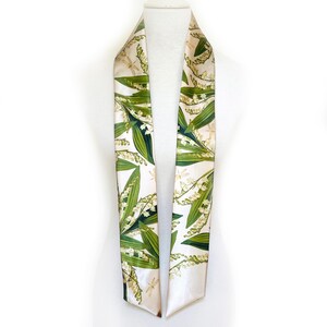 May include: A cream-colored silk scarf featuring a botanical print of lily of the valley flowers and green leaves. The scarf is draped over a mannequin, showcasing its design and texture. The scarf has a white border.