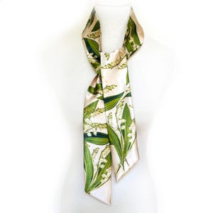 May include: A cream-colored silk scarf featuring a botanical print of lily of the valley flowers and green leaves. The scarf is draped over a white mannequin, showcasing its design and texture. The scarf is a stylish accessory.