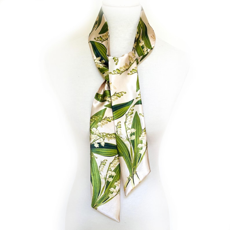 May include: A cream-colored silk scarf featuring a botanical print of lily of the valley flowers and green leaves. The scarf is draped over a white mannequin, showcasing its design and texture. The scarf is a stylish accessory.