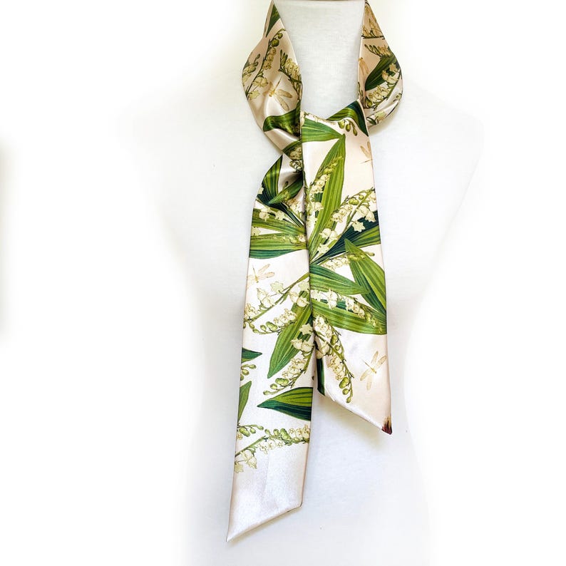 May include: A cream-colored silk scarf with a botanical print of green leaves and white lily of the valley flowers. The scarf is draped over a white mannequin and features a light beige border.