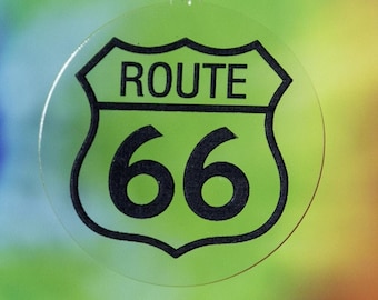 Op de afbeelding: Een ronde, transparante ornament met een zwart-wit Route 66-bord erop gedrukt. De ornament hangt aan een zilveren ketting.