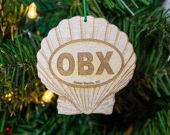 May include: A wooden Christmas ornament shaped like a seashell with the text "OBX Outer Banks, NC" inside. The ornament is hanging on a green Christmas tree.