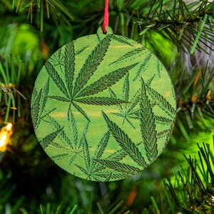 May include: A green Christmas ornament with a pattern of marijuana leaves. The ornament is hanging on a Christmas tree.