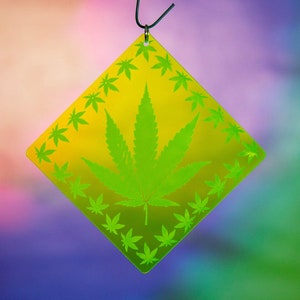 Puede incluir: Un adorno amarillo en forma de diamante con un diseño de hoja de marihuana verde. El adorno está colgado de un gancho plateado.