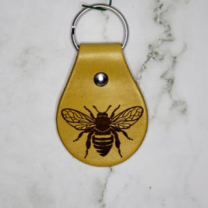 Könnte beinhalten: Ein hellbrauner Leder-Schlüsselanhänger mit einem detaillierten, dunkelbraunen Bienenmuster. Der Schlüsselanhänger hat einen silbernen Metallring und einen Druckknopfverschluss. Die Biene ist mittig auf dem Leder platziert, mit ausgebreiteten Flügeln.