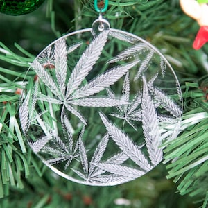 Könnte beinhalten: Ein transparenter Weihnachtsbaumschmuck aus Glas mit einem weißen Marihuana-Blatt-Design. Der Schmuck hängt an einem grünen künstlichen Weihnachtsbaum.