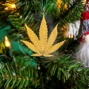 Puede incluir: Un adorno de hoja de marihuana dorada colgado de un árbol de Navidad verde. El adorno está hecho de plástico transparente con un diseño de hoja dorada.