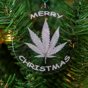 May include: A clear glass Christmas ornament with a white marijuana leaf and the text "Merry Christmas" on a green Christmas tree.