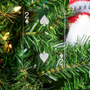 May include: A clear glass ornament shaped like a playing card, featuring the two of spades. The ornament is hanging on a green Christmas tree.