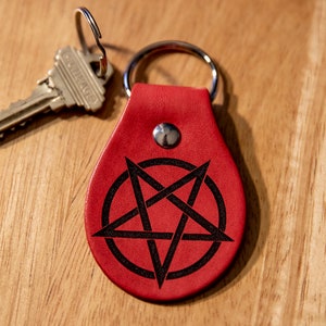 May include: A red leather keychain with a black pentagram design. The keychain has a silver ring and a silver key attached.