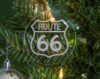 Op de afbeelding: Een transparante glazen kerstbal in de vorm van een Route 66-bord. Het bord is wit en heeft de tekst "Route 66" erop.