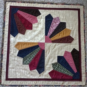 May include: A square quilt with a beige background and a multi-colored design. The design features four fan-shaped patterns in shades of red, pink, brown, gold, green, blue, and black. The quilt is framed with a brown and white border.
