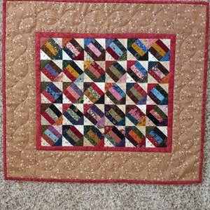 May include: A small quilt with a red border and a brown background. The quilt is made up of many small squares of different fabrics, each with a different pattern. The quilt is quilted with a simple pattern of small circles.