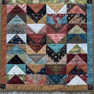 May include: A handcrafted quilt featuring a patchwork design with various fabrics in shades of brown, blue, and red. The quilt has a geometric pattern with triangle shapes and a decorative border. The quilt is square and appears to be machine-stitched.