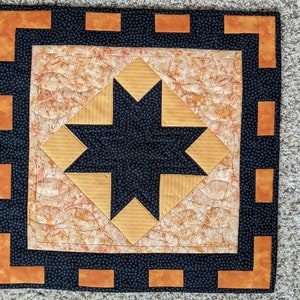 May include: A square quilted textile with a central eight-pointed star design in black, surrounded by orange and tan geometric shapes. The border is black with orange corner accents. The quilt is likely handmade.