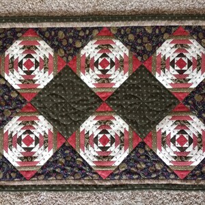 May include: A quilted table runner with a geometric pattern of pineapples in shades of red, white, and green. The runner has a dark green background with a floral print and a brown border.