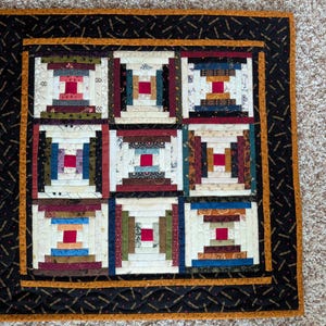 May include: A square fabric quilt featuring a nine-block log cabin pattern. Each block has a central red square surrounded by strips of various colors and patterns, including cream, blue, green, and brown. The quilt has a black and gold border.