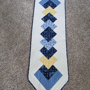 May include: A quilted table runner with a diamond pattern. The runner features a cream-colored background with blue and yellow diamond shapes. The diamonds are made of patterned fabric and are arranged in a vertical line. The edges are finished with a blue border.