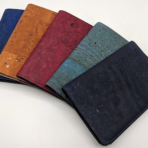 May include: Five cork wallets in different colors: blue, brown, red, teal, and black. The wallets are arranged in a fan shape, with the blue wallet on the left and the black wallet on the right.
