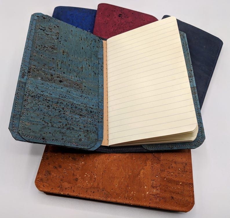 May include: A blue cork notebook with lined pages, open to reveal the blank pages. The notebook is surrounded by other cork notebooks in different colors.