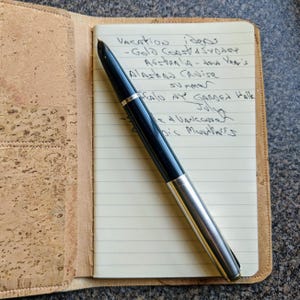 May include: A close-up of a notebook with a list of vacation ideas written in black ink. The notebook is open and a black and silver fountain pen is lying on top of it. The notebook is sitting on a cork surface.