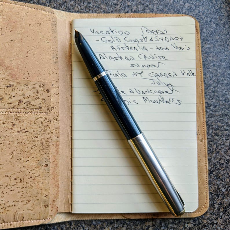 May include: A close-up of a notebook with a list of vacation ideas written in black ink. The notebook is open and a black and silver fountain pen is lying on top of it. The notebook is sitting on a cork surface.
