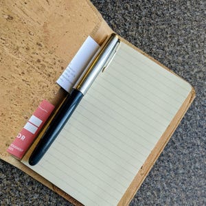 May include: A cork notebook with a lined page and a silver and black pen. The notebook has a red bookmark with the word "OR" and the text "throughout 2020".