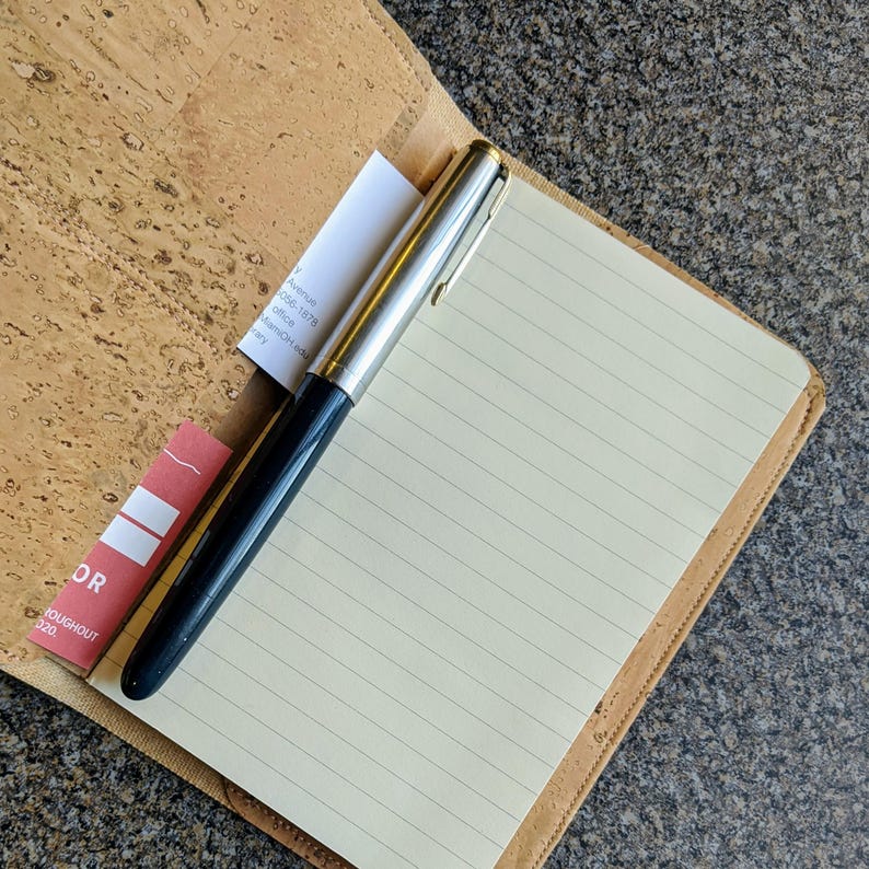 May include: A cork notebook with a lined page and a silver and black pen. The notebook has a red bookmark with the word "OR" and the text "throughout 2020".