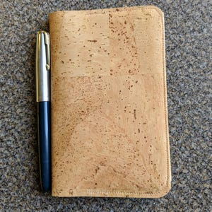 May include: A cork notebook cover with a pen next to it. The cover is a light brown color with a subtle pattern of cork.
