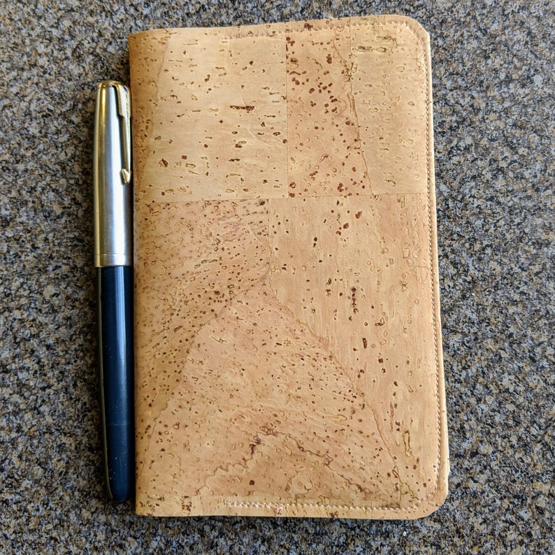 May include: A cork notebook cover with a pen next to it. The cover is a light brown color with a subtle pattern of cork.