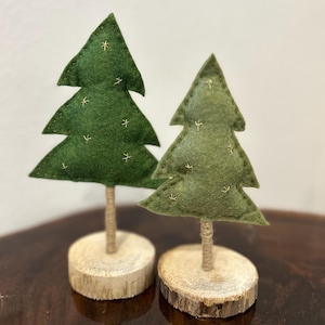 Puede incluir: Dos árboles de Navidad decorativos de fieltro. Los árboles son verdes con detalles de estrellas doradas y están unidos a pequeñas bases de madera. Los troncos de los árboles están envueltos en hilo. Los árboles están sobre una superficie marrón oscuro.
