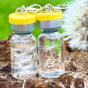 May include: Two clear glass vials with yellow tops and silver caps, each containing a single dandelion seed head. The vials are attached to silver chains with clasps.