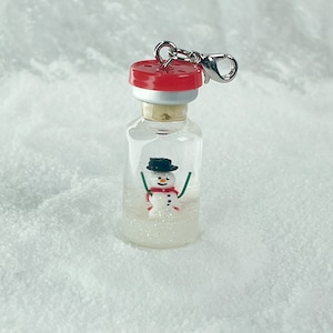 Puede incluir: Un pequeño charm de botella de vidrio con un muñeco de nieve en su interior. La botella tiene un tapón de corcho rojo y blanco y un cierre plateado. El muñeco de nieve lleva un sombrero negro y una bufanda roja.