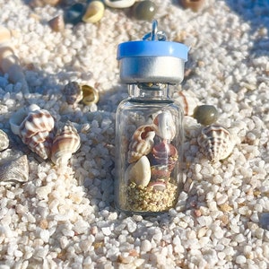 May include: A small glass bottle filled with seashells and sand, with a blue and silver lid. The bottle is sitting on a bed of white sand.