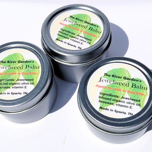 May include: Three small, silver tins of Jewelweed Balm. Each tin has a white label with green leaf illustrations and text that reads "The River Garden's Jewelweed Balm" and "Apply to rash or bug bite."
