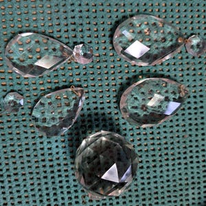 May include: A collection of clear, faceted crystal chandelier prisms in various shapes and sizes. The prisms include teardrop, oval, and round shapes, with small metal loops for hanging. The crystals reflect light, creating a sparkling effect.