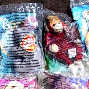 May include: Two Ty Teenie Beanie Babies plush toys sealed in clear plastic bags. One is a grey animal with a heart-shaped Ty tag, and the other is a red monkey with a tan face and ears. Both bags feature printed safety warnings and text.