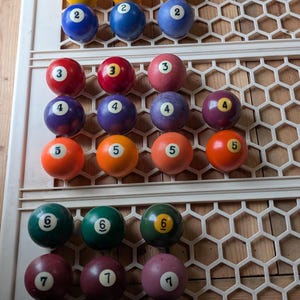May include: A collection of thirty-two billiard balls arranged in rows on a white plastic grid. The set includes white cue balls, yellow ones, blue twos, red threes, purple fours, orange fives, green sixes, maroon sevens, and black eights.