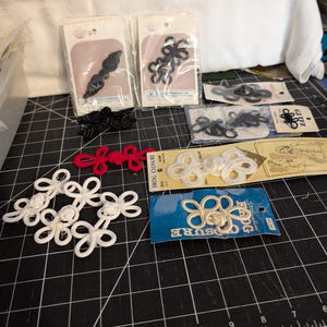 May include: A collection of assorted frog closures in black, white, and red, presented on various branded cardboard backing cards and loose, featuring intricate knotted cord designs for garment fastening, arranged on a black cutting mat.