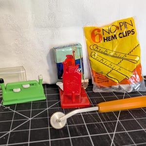 May include: A collection of sewing tools, including a green seam ripper, a red rotary cutter, a package of yellow hem clips, and a tracing wheel with an orange handle. The tools are arranged on a black grid surface.