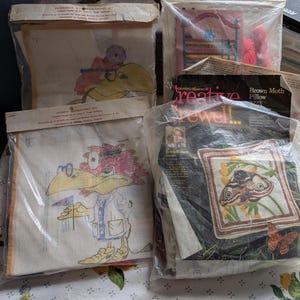 May include: A collection of four needlepoint and crewel embroidery kits in clear plastic packaging. Two kits feature a cartoon bird character, one shows a bedroom scene, and one displays a brown moth design with the text Creative Crewel.